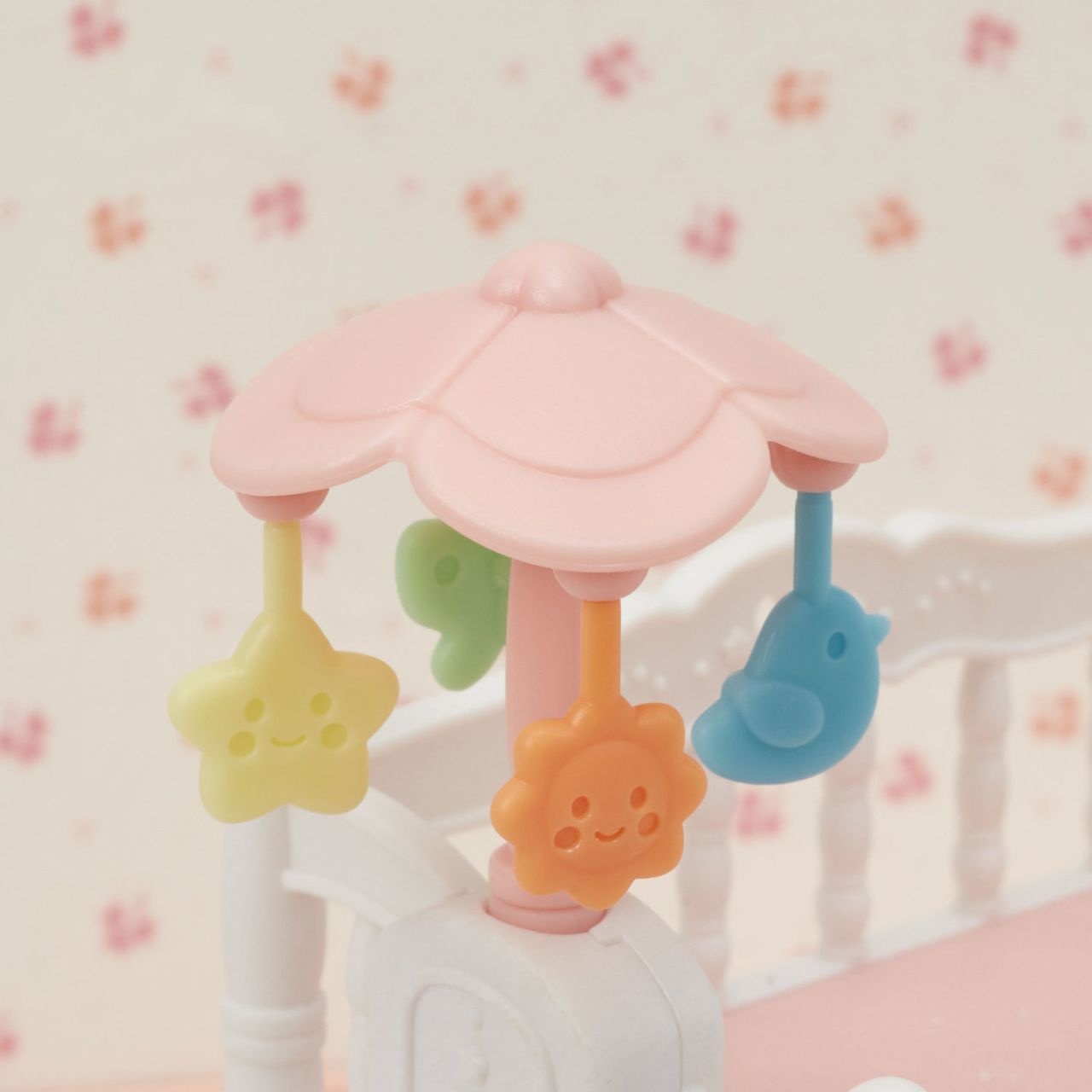 A Calico Critters Crib with Mobile featuring a pink crib mobile adorned with yellow star, orange flower, and blue bird-shaped hanging toys, displayed above a white crib against a floral wallpaper background—ideal for completing your whimsical baby nursery set or pairing with other Calico Critters dollhouse furniture.