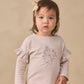 A toddler with brown hair is wearing the Quincy Mae Baby Girls’ and Girls’ Ruffle Long Sleeve Tee, an everyday essential adorned with floral details and ruffled sleeves, paired with a bow hair accessory, standing against a plain beige background.