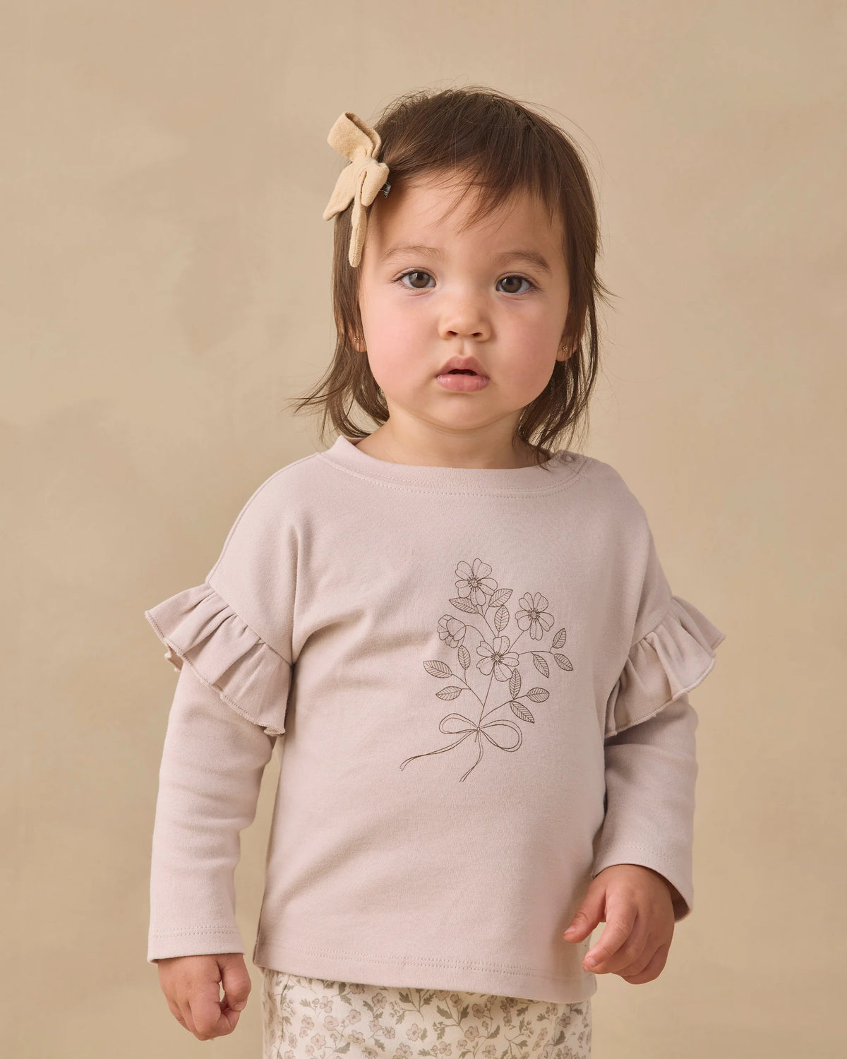 A toddler with brown hair is wearing the Quincy Mae Baby Girls’ and Girls’ Ruffle Long Sleeve Tee, an everyday essential adorned with floral details and ruffled sleeves, paired with a bow hair accessory, standing against a plain beige background.