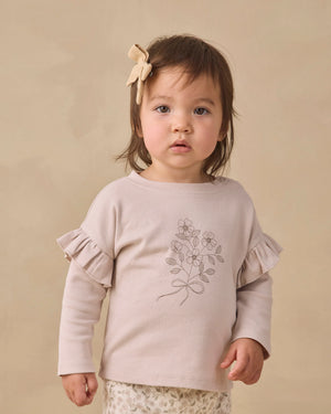 A toddler with brown hair is wearing the Quincy Mae Baby Girls’ and Girls’ Ruffle Long Sleeve Tee, an everyday essential adorned with floral details and ruffled sleeves, paired with a bow hair accessory, standing against a plain beige background.