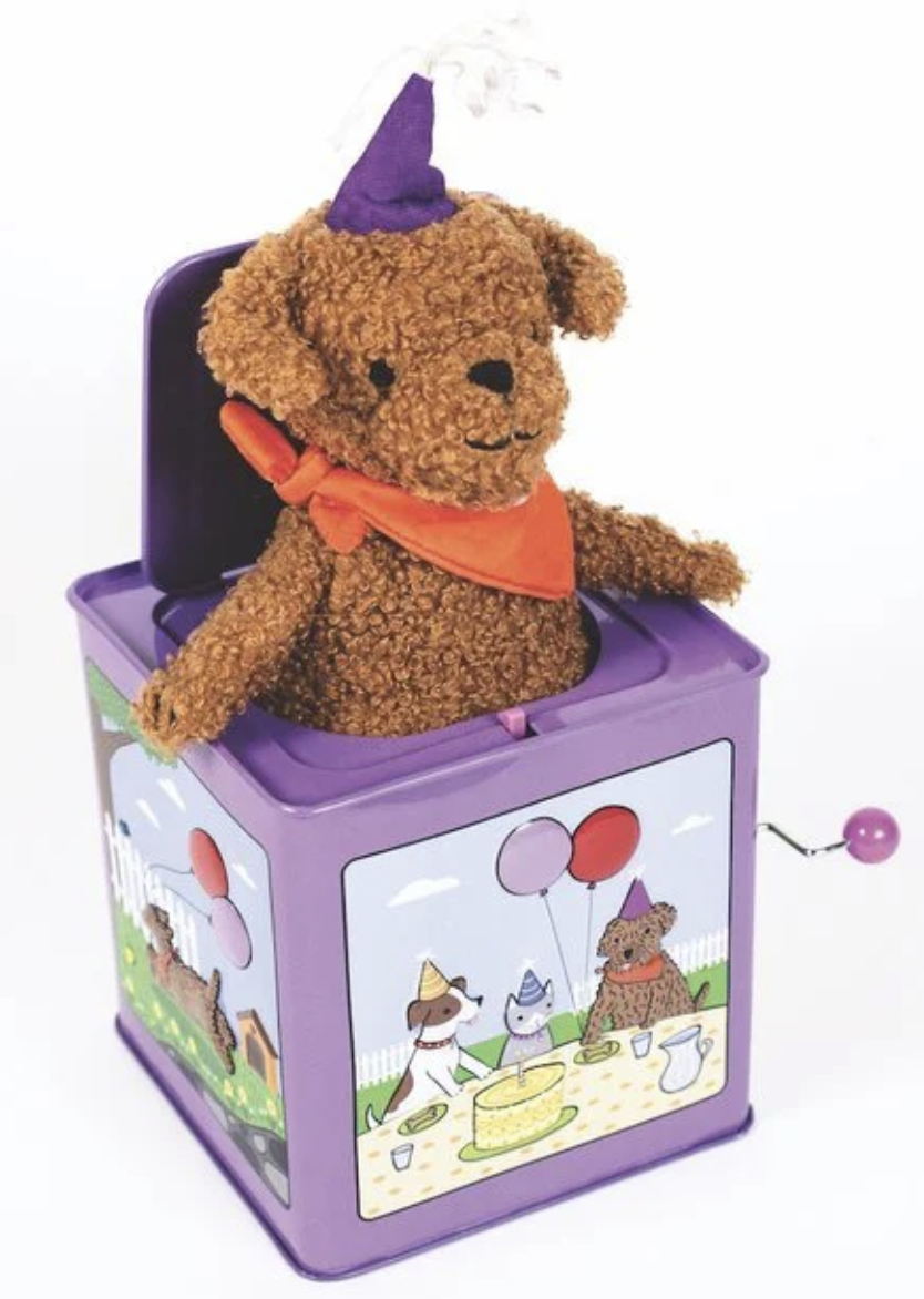 A charming plush brown teddy bear wearing a purple hat and orange scarf pops out of the Jack Rabbit Creations Jack-in-the-Box, a whimsical purple toy adorned with festive party scenes—a delightful homage to classic jack-in-the-box toys.