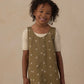 A smiling young child with curly hair wears the Rylee & Cru Girls’ V-Neck Overall Dress—an olive green piece adorned with white flower patterns and front patch pockets—layered over a short-sleeved cream shirt, standing against a plain beige background.