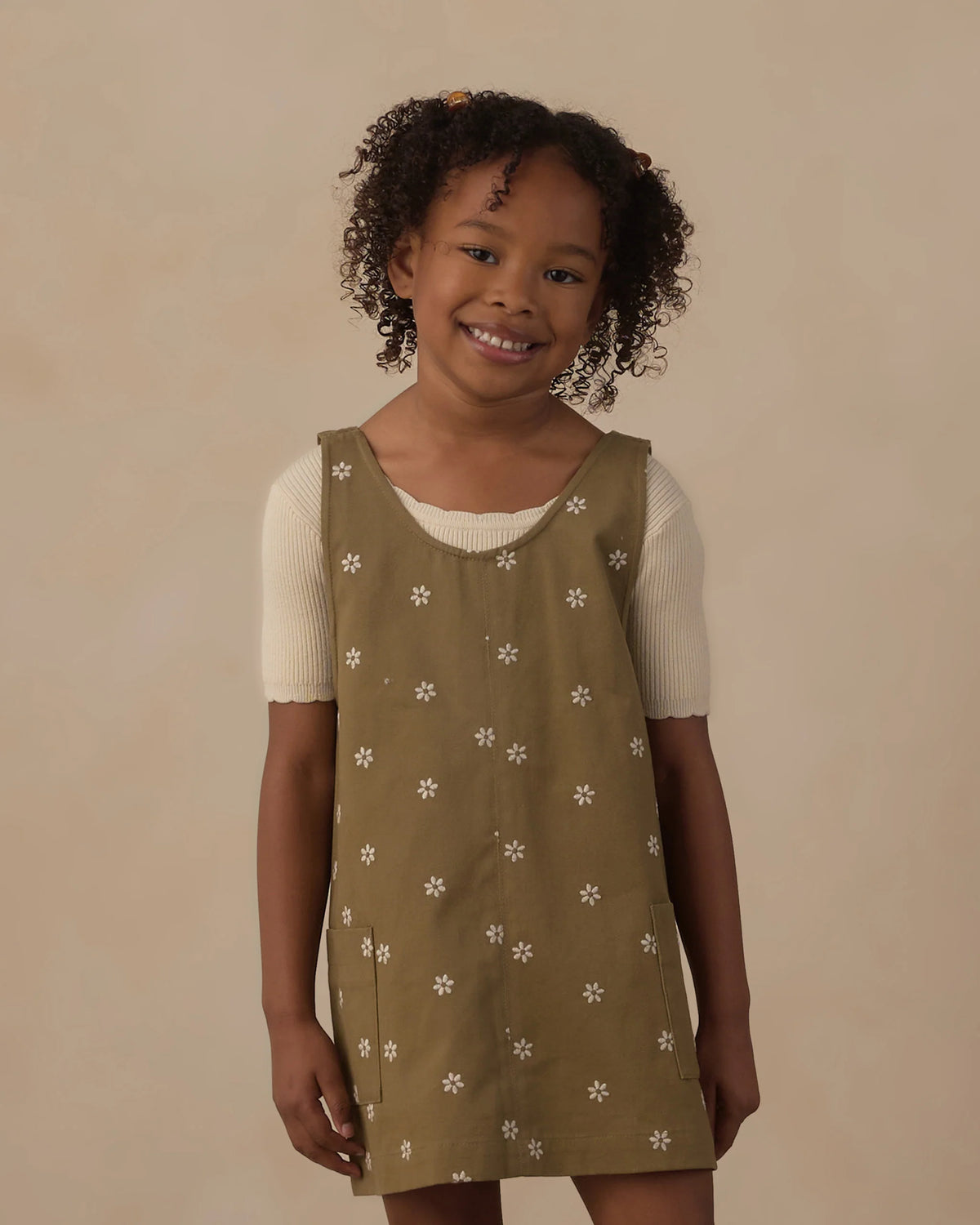 A smiling young child with curly hair wears the Rylee & Cru Girls’ V-Neck Overall Dress—an olive green piece adorned with white flower patterns and front patch pockets—layered over a short-sleeved cream shirt, standing against a plain beige background.