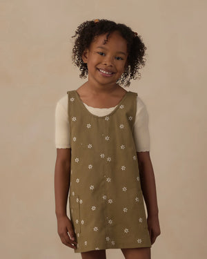 A smiling young child with curly hair wears the Rylee & Cru Girls’ V-Neck Overall Dress—an olive green piece adorned with white flower patterns and front patch pockets—layered over a short-sleeved cream shirt, standing against a plain beige background.