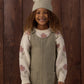 A smiling child with curly hair stands in front of a wooden wall, wearing a knit hat, a cream sweater with pink flowers, and the Rylee & Cru Girls’ Odette Overall Dress in khaki denim with front pockets.