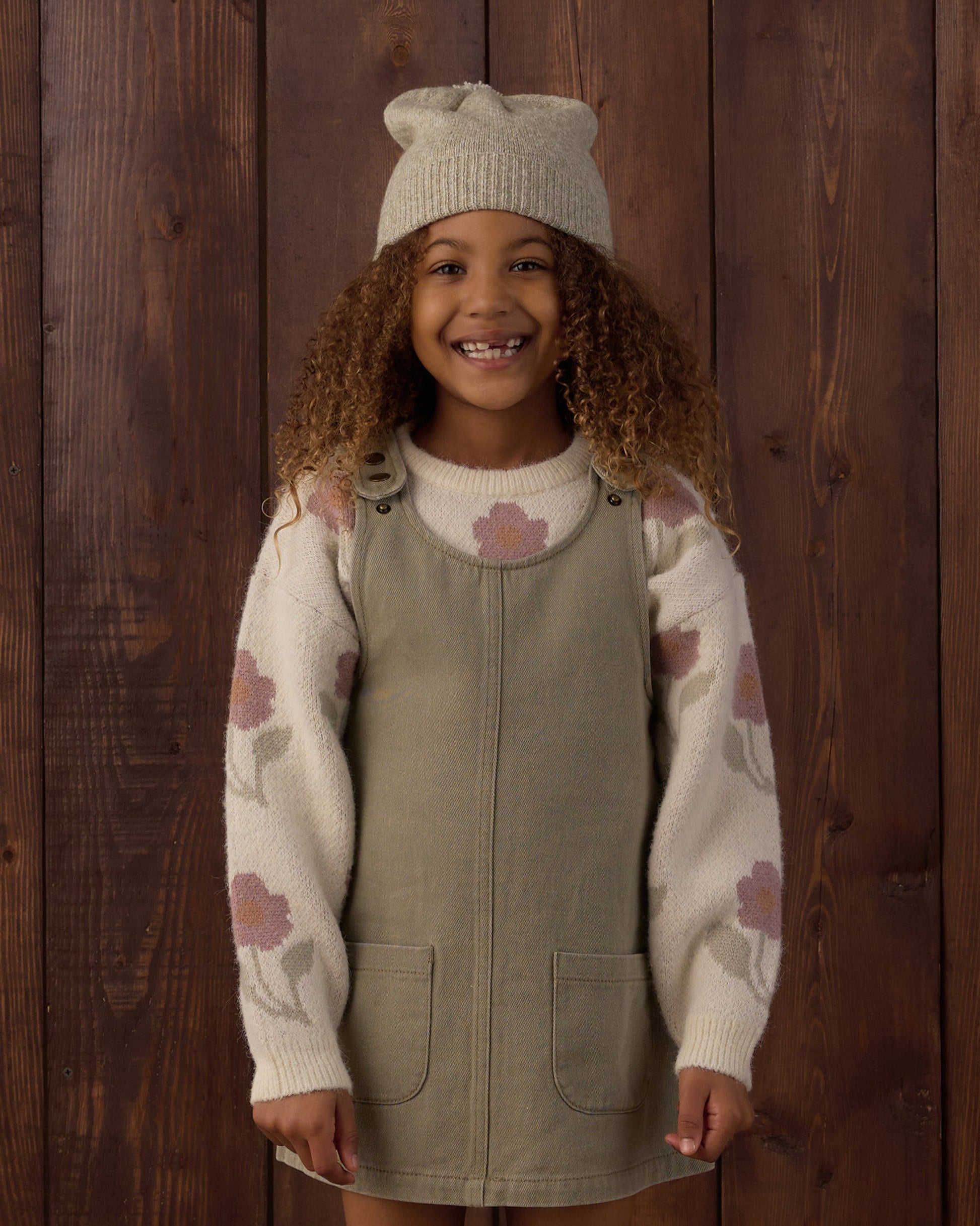A smiling child with curly hair stands in front of a wooden wall, wearing a knit hat, a cream sweater with pink flowers, and the Rylee & Cru Girls’ Odette Overall Dress in khaki denim with front pockets.