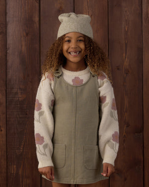 A smiling child with curly hair stands in front of a wooden wall, wearing a knit hat, a cream sweater with pink flowers, and the Rylee & Cru Girls’ Odette Overall Dress in khaki denim with front pockets.