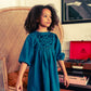 A young girl with curly hair stands indoors wearing the Louis Louise Girls' Mahana Light Denim Dress, with a wicker chair, red record player, and stacked books in the background.