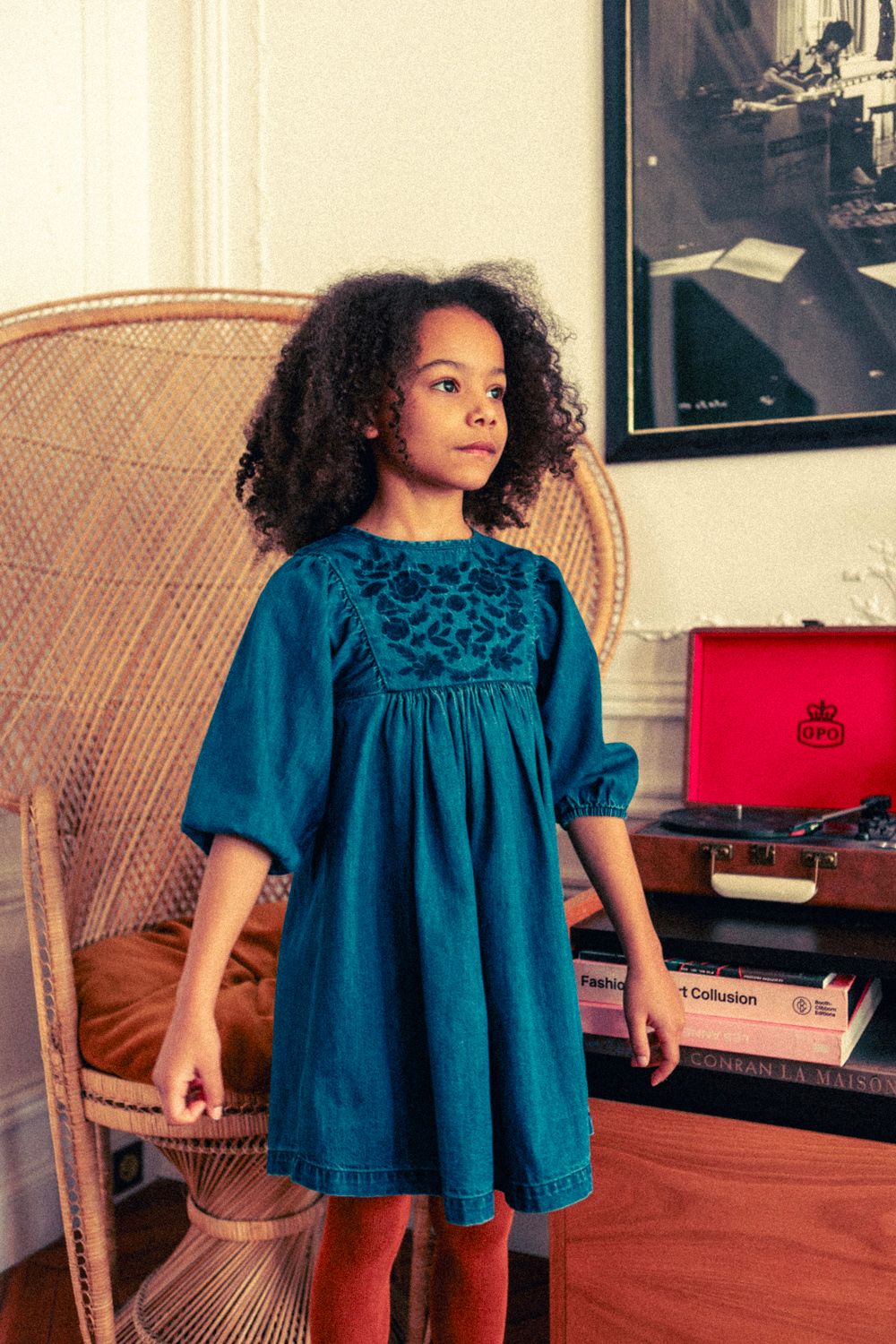 A young girl with curly hair stands indoors wearing the Louis Louise Girls' Mahana Light Denim Dress, with a wicker chair, red record player, and stacked books in the background.