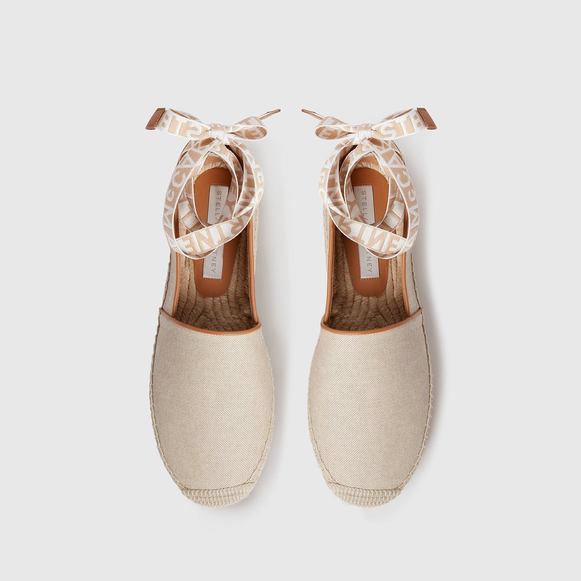 A pair of beige Stella McCartney Gaia Canvas Flat sandals with textured canvas, woven soles, and ribbon ankle ties, shown top-down on a white background. Lightweight and stylish.