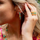 A woman wears Juliska Capucine De Wulf Berry Gem Medium Hoop Earrings with turquoise stones, along with matching rings and a necklace, while holding her hand near her ear.