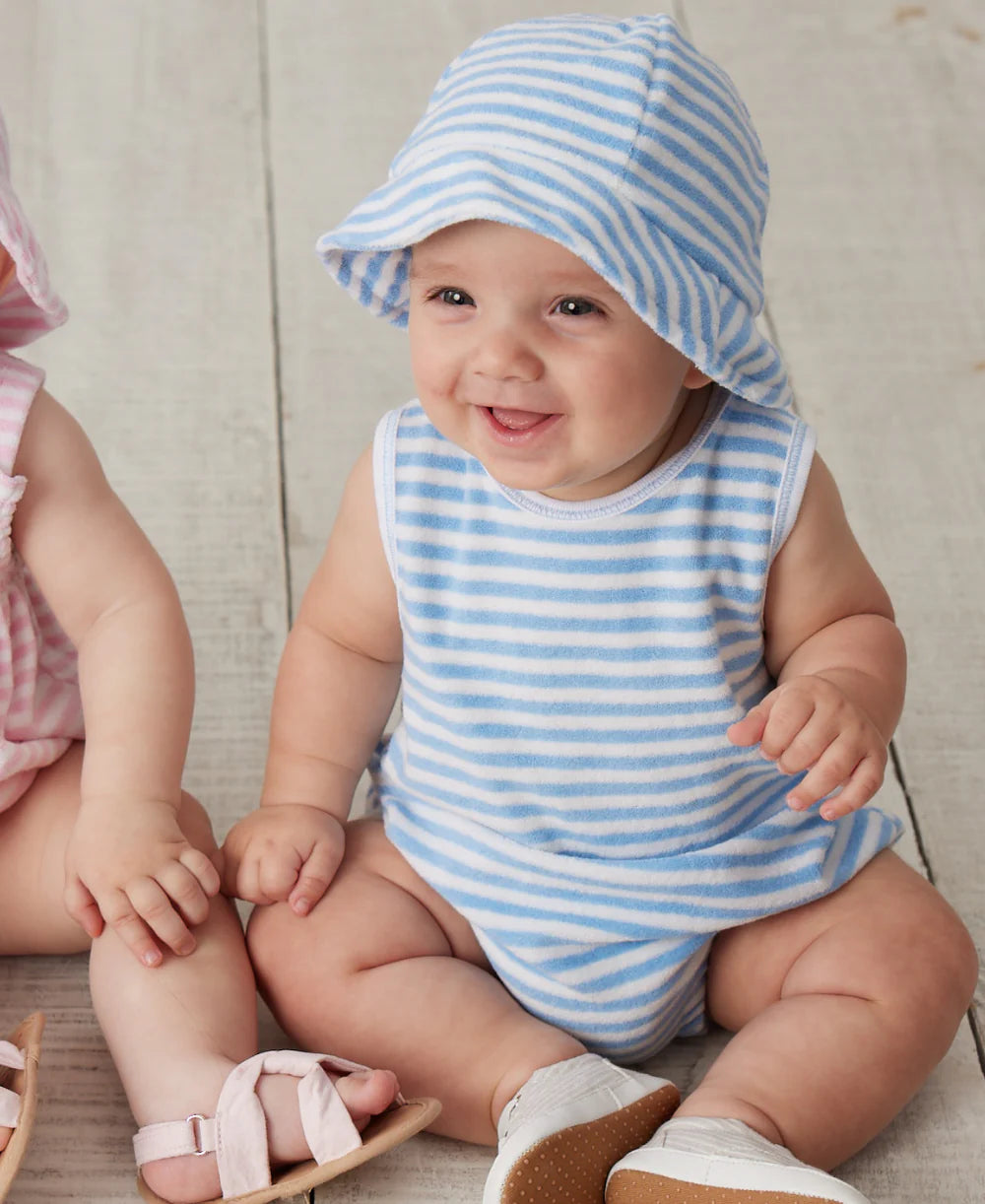 A baby smiles while wearing the Kissy Kissy Baby Cabana Terry Stripes Sun Hat with a matching blue and white romper, sitting on a wooden floor. Another baby in pink is partially visible beside them.