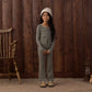 A young girl stands indoors on a rug, surrounded by wooden walls and vintage furniture, wearing the Rylee & Cru Girls’ Flare Rib Knit Pant with an elastic waistband, paired with a striped pajama top and topped with a cozy knit hat.