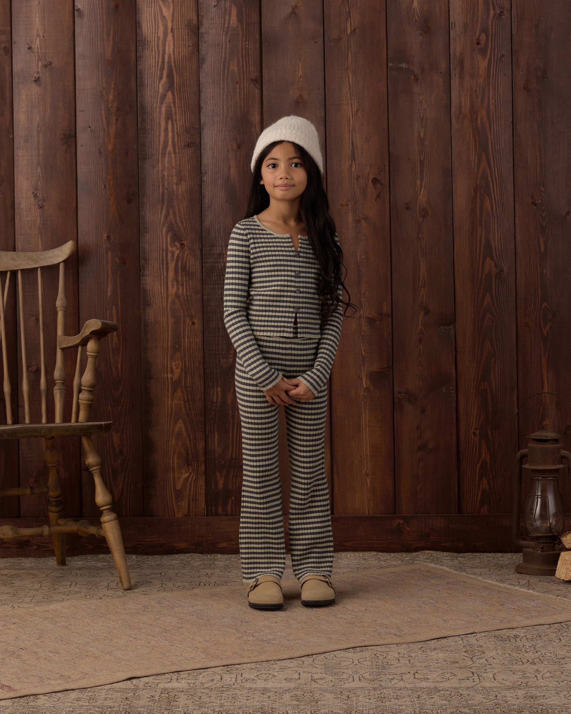 A young girl stands indoors on a rug, surrounded by wooden walls and vintage furniture, wearing the Rylee & Cru Girls’ Flare Rib Knit Pant with an elastic waistband, paired with a striped pajama top and topped with a cozy knit hat.