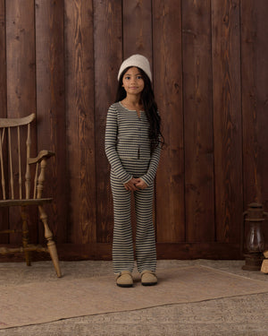 A young girl stands indoors on a rug, surrounded by wooden walls and vintage furniture, wearing the Rylee & Cru Girls’ Flare Rib Knit Pant with an elastic waistband, paired with a striped pajama top and topped with a cozy knit hat.
