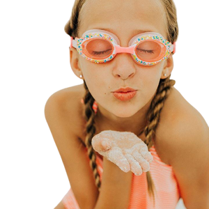 Girl wearing Bling2O Cake Pop Swim Goggles, Angel Cake Pink with pink frames and silicone strap, blowing sand off her hand.