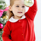 A young girl in a Richmond Red Baby Girls’ and Girls’ Cambridge Cardigan with pearl buttons by The Beaufort Bonnet Company holds up a gold Christmas ornament next to a decorated tree.