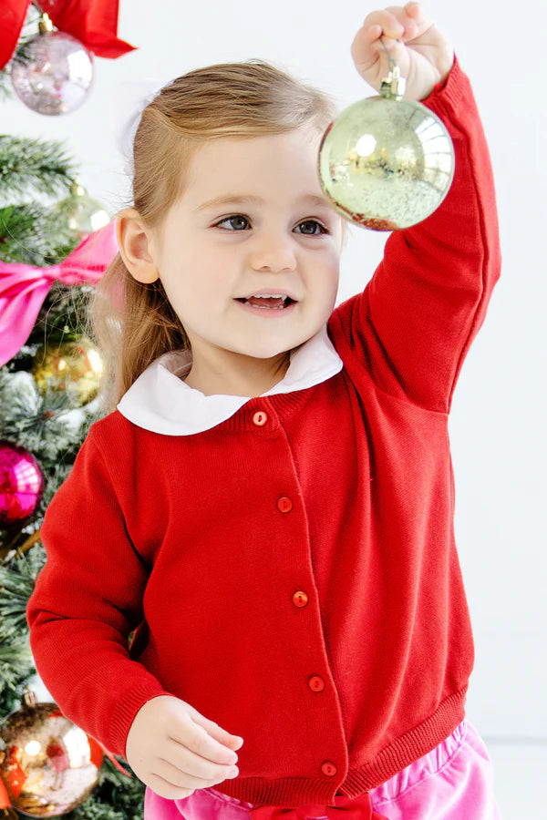 A young girl in a Richmond Red Baby Girls’ and Girls’ Cambridge Cardigan with pearl buttons by The Beaufort Bonnet Company holds up a gold Christmas ornament next to a decorated tree.