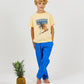 A young boy wearing a relaxed fit, yellow Arsene & Les Pipelettes Boys' and Girls' Caribbean T-Shirt and blue pants stands barefoot next to a pineapple and two oranges against a plain white background.