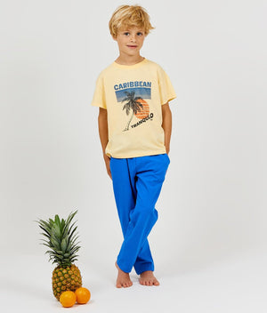 A young boy wearing a relaxed fit, yellow Arsene & Les Pipelettes Boys' and Girls' Caribbean T-Shirt and blue pants stands barefoot next to a pineapple and two oranges against a plain white background.