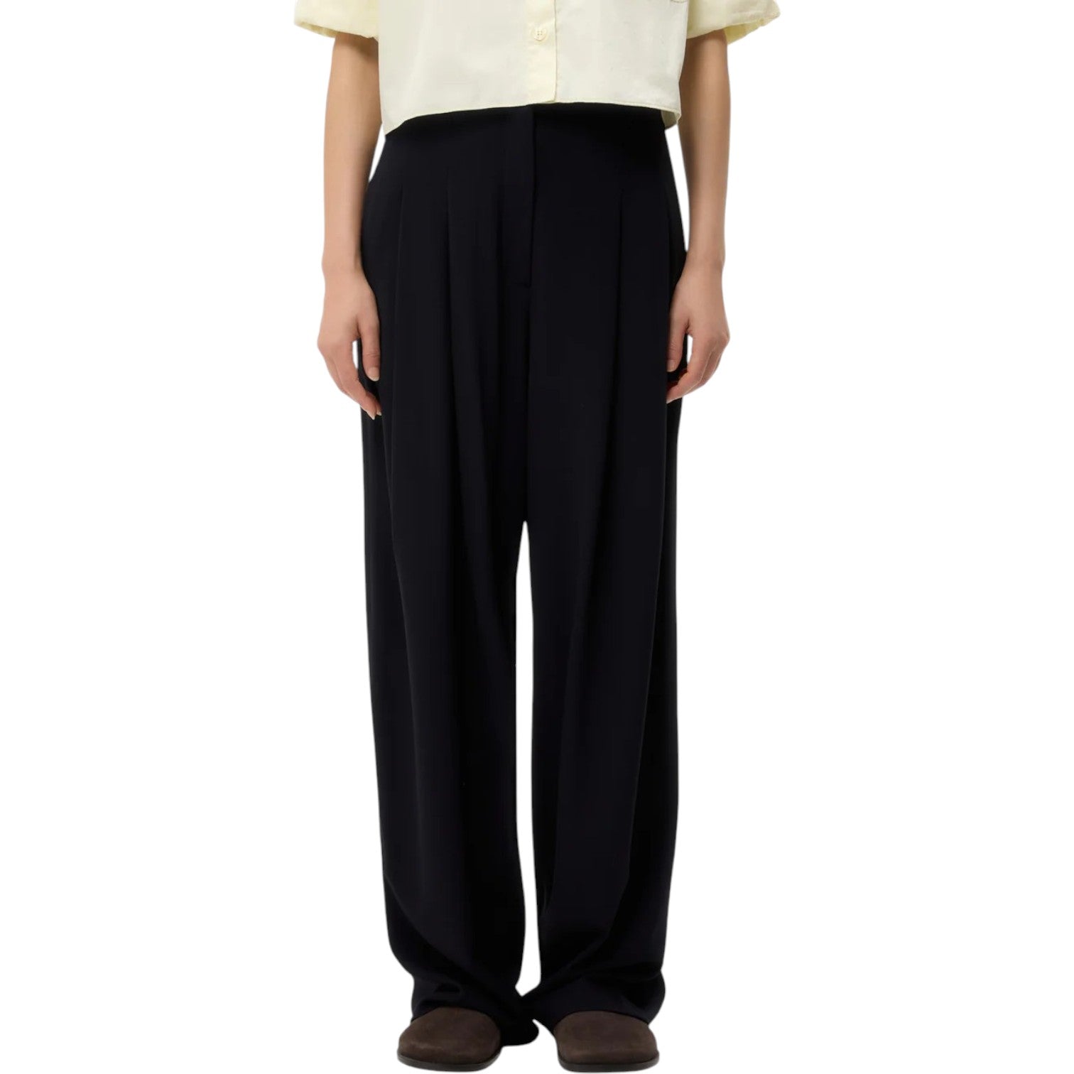Wearing a light yellow short-sleeve shirt, TWP Carries Trouser in black, and dark shoes, a person stands facing forward against a white background.