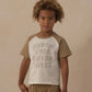 A young child stands against a beige background wearing the Rylee & Cru Baby Boys’ and Boys’ Raglan Short Sleeve Tee crafted from soft cotton jersey, featuring “ADVENTURE ENTHUSIAST” across the front, paired with matching shorts.