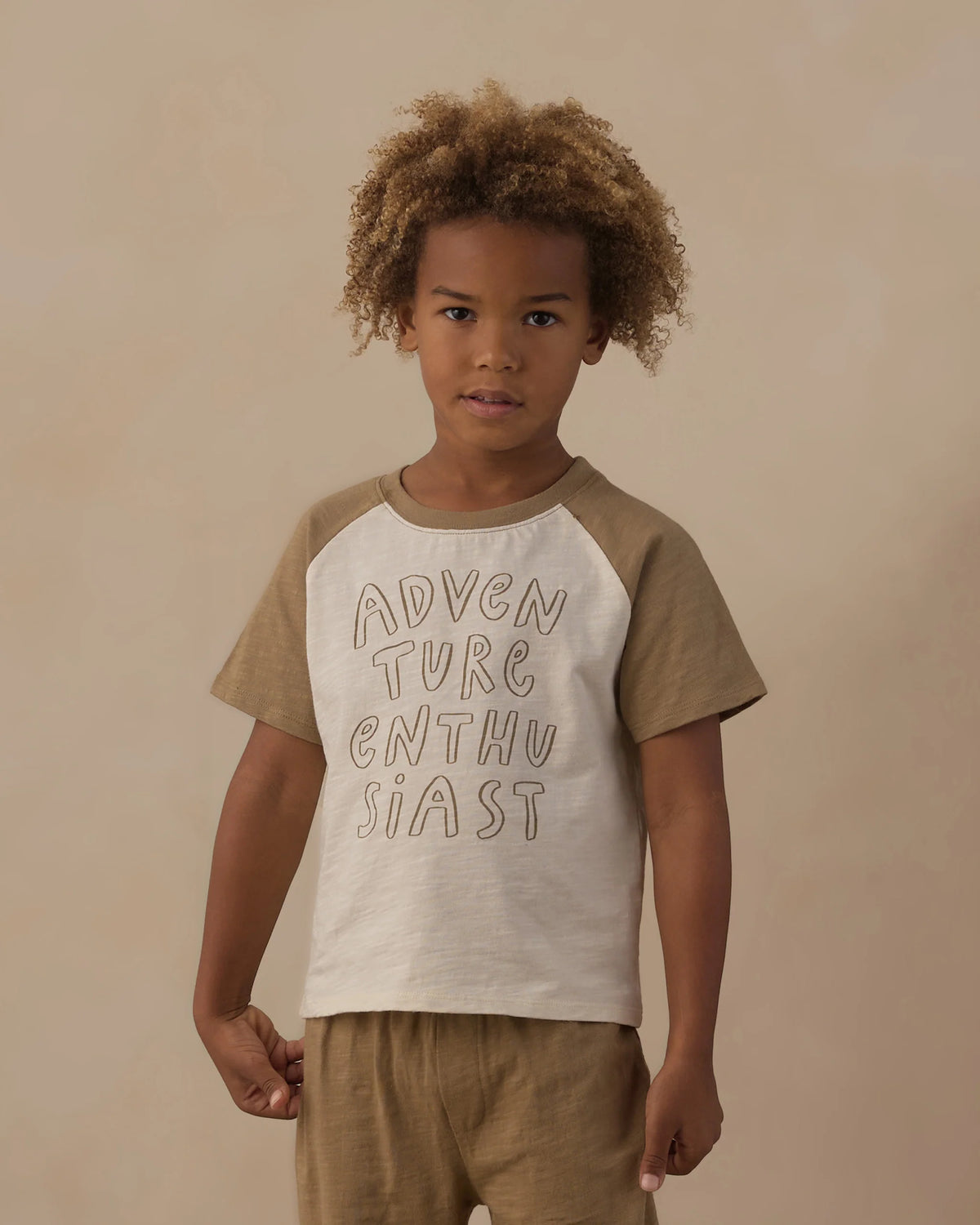 A young child stands against a beige background wearing the Rylee & Cru Baby Boys’ and Boys’ Raglan Short Sleeve Tee crafted from soft cotton jersey, featuring “ADVENTURE ENTHUSIAST” across the front, paired with matching shorts.