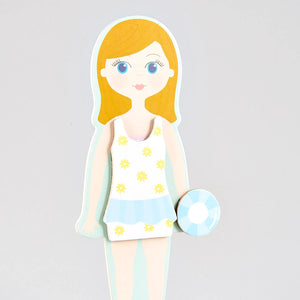 Illustration of the Elsie Magnetic Dress Up Character by Floss and Rock—a girl with blonde hair in a white and yellow floral swimsuit, holding a blue and white float, standing against a plain gray background—reminiscent of a magnetic dress up doll ready to showcase her summer style.