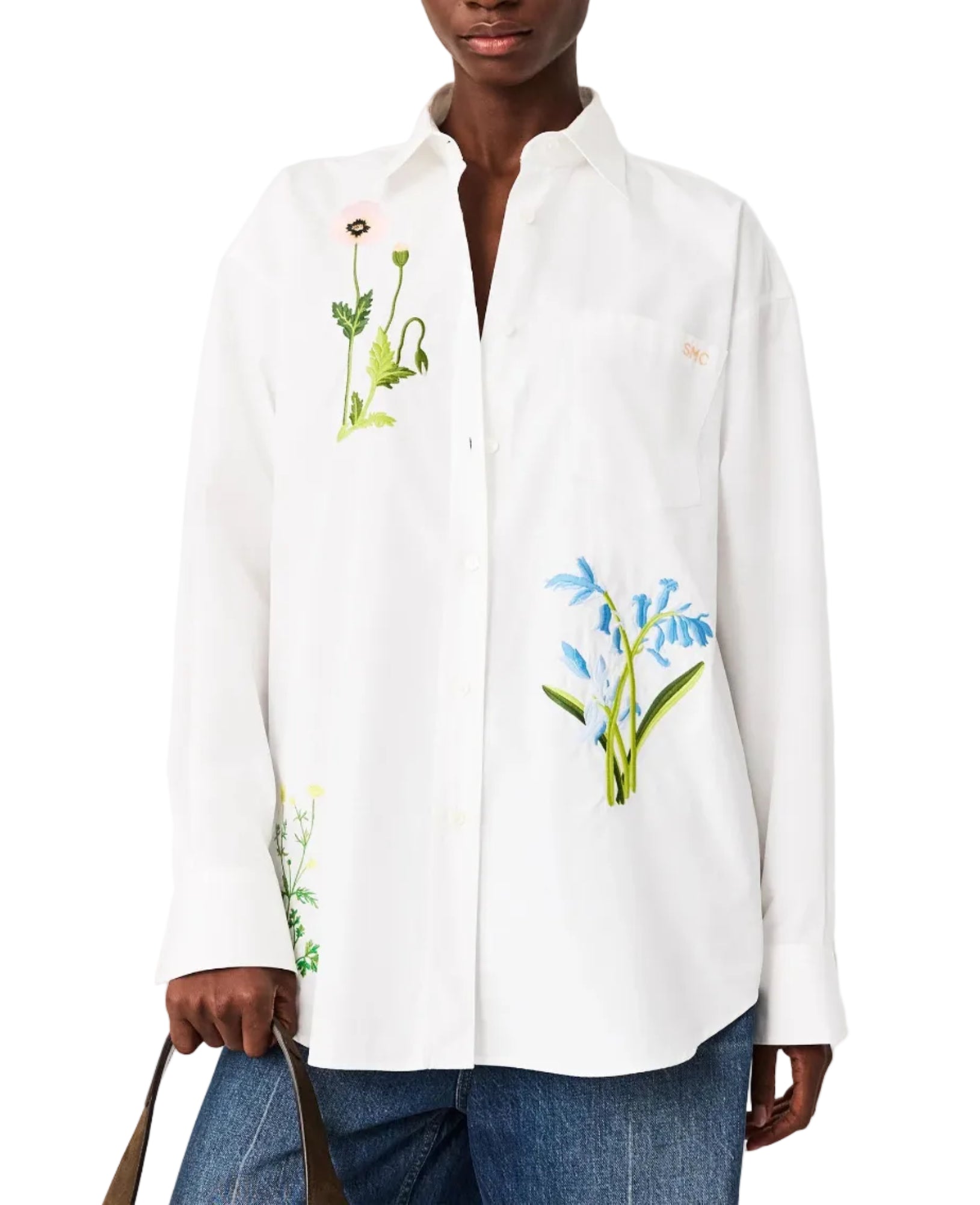 A person wears the Stella McCartney Flower Embroidered Shirt, a loose boyfriend-fit white button-up with botanical designs, paired with blue jeans and carrying a brown strap bag.