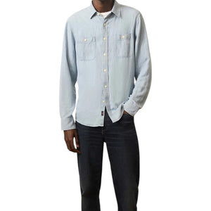 A person wears the Faherty Tried and True Chambray Workshirt in light blue, featuring two chest pockets, paired with dark jeans and standing with one hand in a pocket.