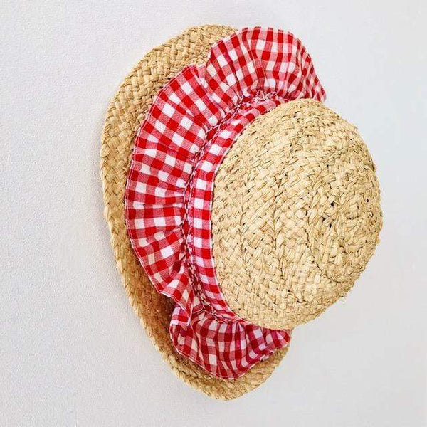 The Girls' Nathalie Smocked Raffia Hat from Kidiwi, featuring a red and white checkered fabric ruffle, is hanging on a white wall.