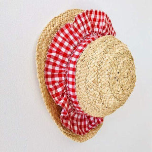 The Girls' Nathalie Smocked Raffia Hat from Kidiwi, featuring a red and white checkered fabric ruffle, is hanging on a white wall.
