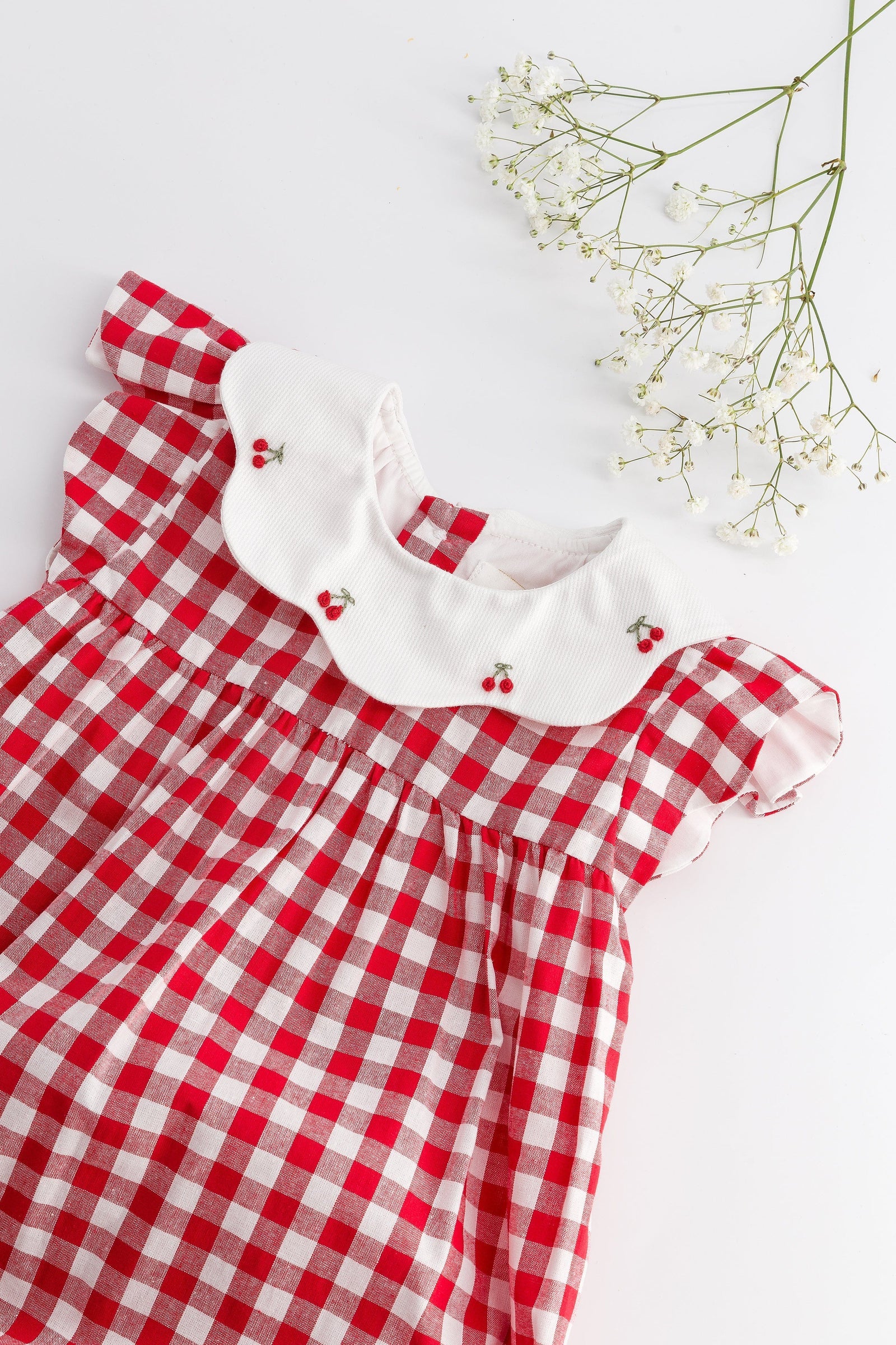The Kidiwi Baby Girls' Charlotte Romper features red and white gingham with a white scalloped collar and embroidered cherries, shown alongside baby's breath flowers on a white background.