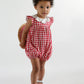 A toddler in the Kidiwi Baby Girls' Charlotte Romper, a red gingham cotton suit with a white collar, pairs it with white sandals and holds a straw hat behind her back against a plain background.