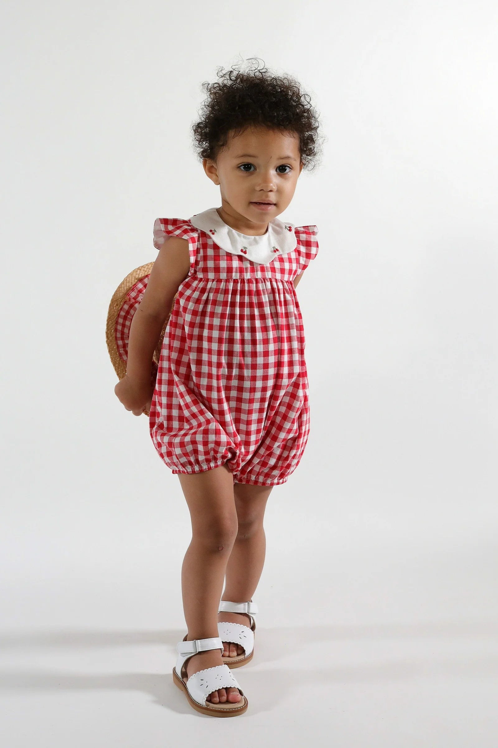 A toddler in the Kidiwi Baby Girls' Charlotte Romper, a red gingham cotton suit with a white collar, pairs it with white sandals and holds a straw hat behind her back against a plain background.