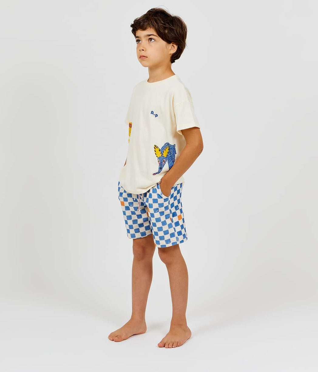 A barefoot child, hands in pockets, stands against a plain light background wearing a cream graphic T-shirt paired with Arsene & Les Pipelettes Boys' Checkered Terry Bermuda Shorts.