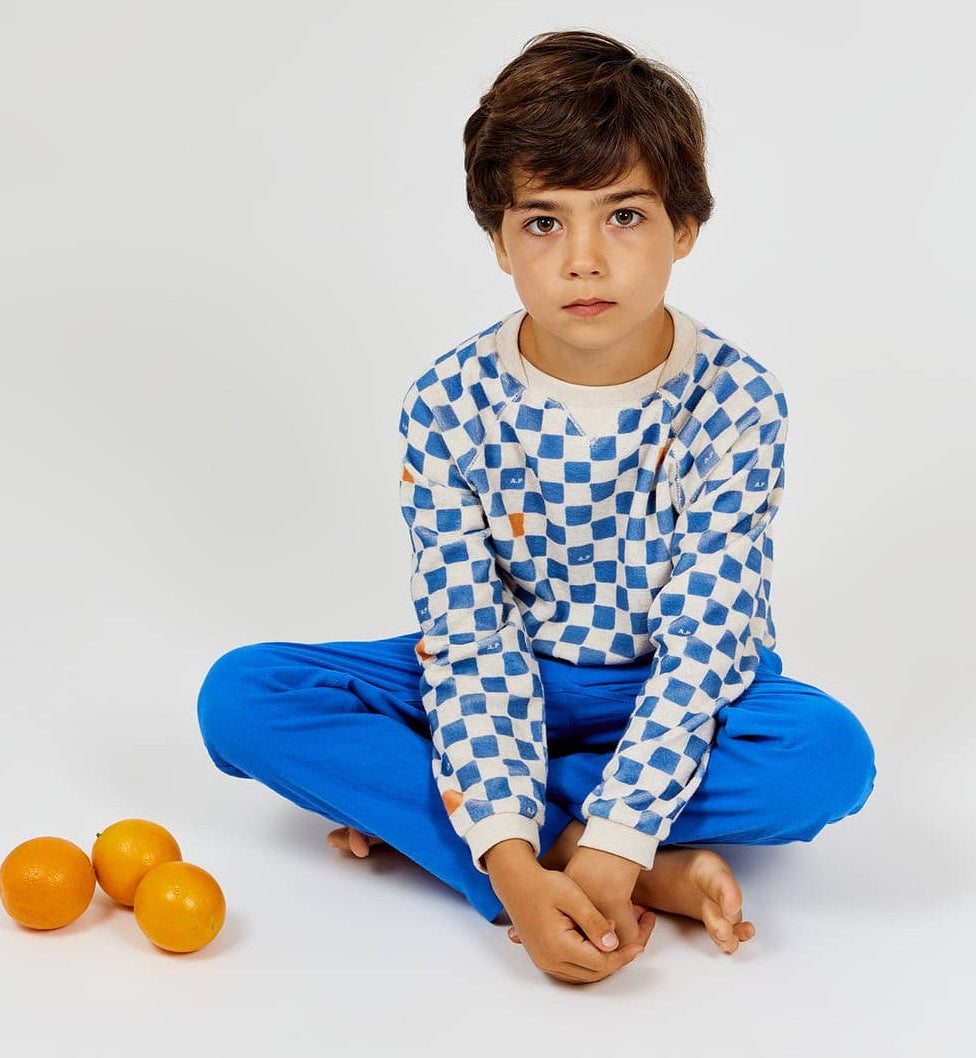 A boy sits cross-legged on the floor wearing the Arsene & Les Pipelettes Boys' Checkered Terry Cloth Sweatshirt and blue pants, with three oranges placed beside him on a white background.