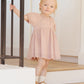 A young child in a pink Quincy Mae Baby Girls' and Girls' Bella Dress stands on wooden stairs, holding a black railing and looking forward with a neutral expression.