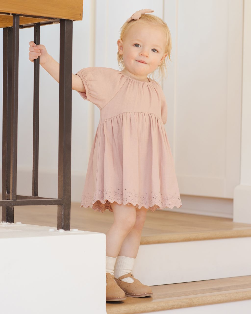 A young child in a pink Quincy Mae Baby Girls' and Girls' Bella Dress stands on wooden stairs, holding a black railing and looking forward with a neutral expression.