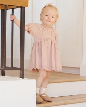 A young child in a pink Quincy Mae Baby Girls' and Girls' Bella Dress stands on wooden stairs, holding a black railing and looking forward with a neutral expression.