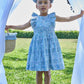 A young girl stands on grass in the Kidiwi Baby Girls' and Girls' Clarisse Smocked Dress, wearing white sandals and holding open white curtains outdoors on a sunny day.