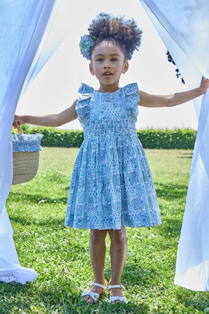 A young girl stands on grass in the Kidiwi Baby Girls' and Girls' Clarisse Smocked Dress, wearing white sandals and holding open white curtains outdoors on a sunny day.