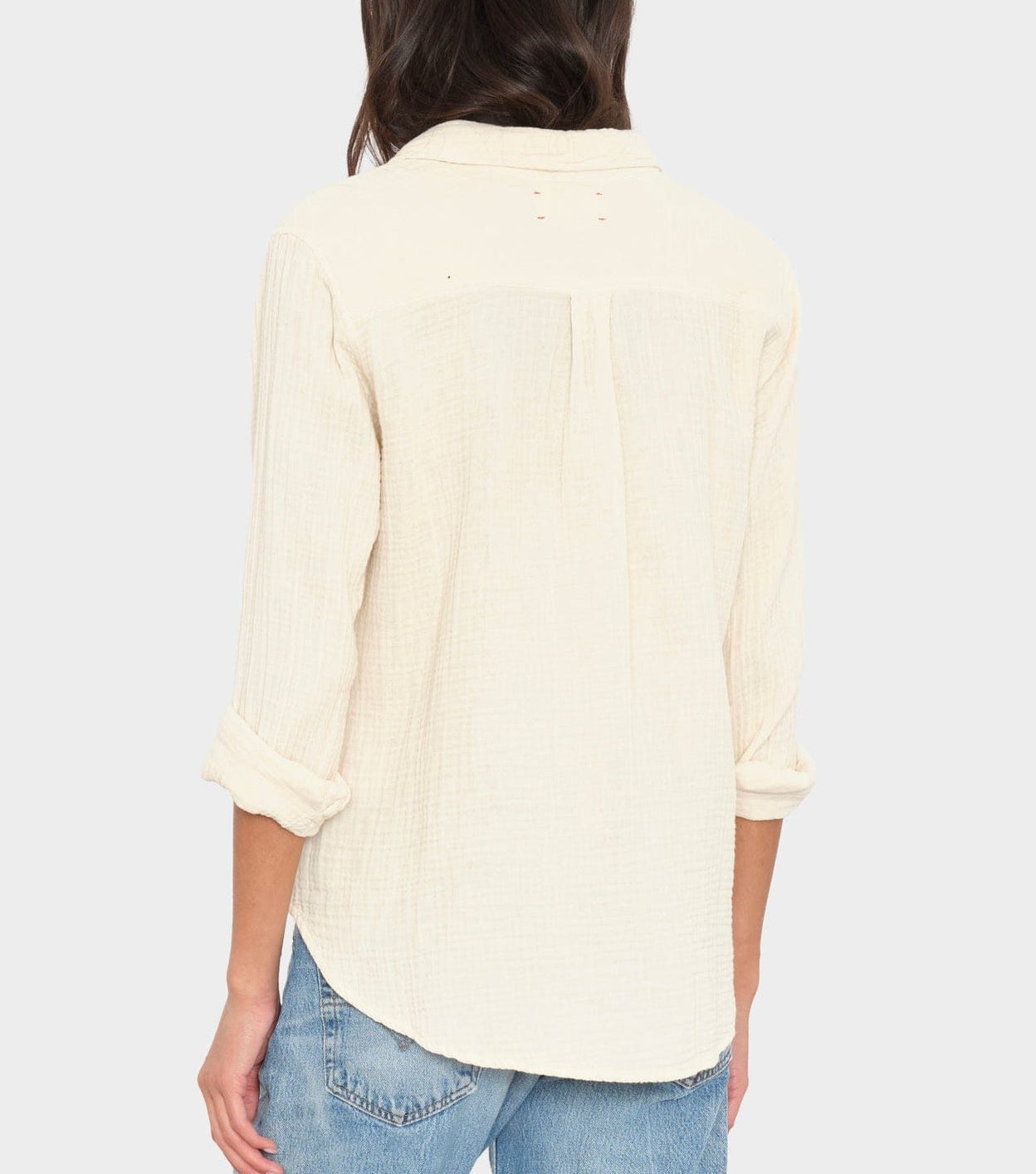 A person is seen from the back wearing the Xirena Scout Cotton Gauze Shirt in cream, featuring rolled cuffs and a shirttail hem, paired with light blue jeans.