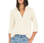 A woman wears the Xirena Scout Cotton Gauze Shirt in cream, featuring a lightweight button-front design, shirttail hem, and rolled sleeves, paired with light blue jeans against a plain background.