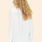 A person with long curly blonde hair, seen from behind against a beige background, wears Xirena's Scout Shirt in soft white cotton gauze paired with light blue jeans.