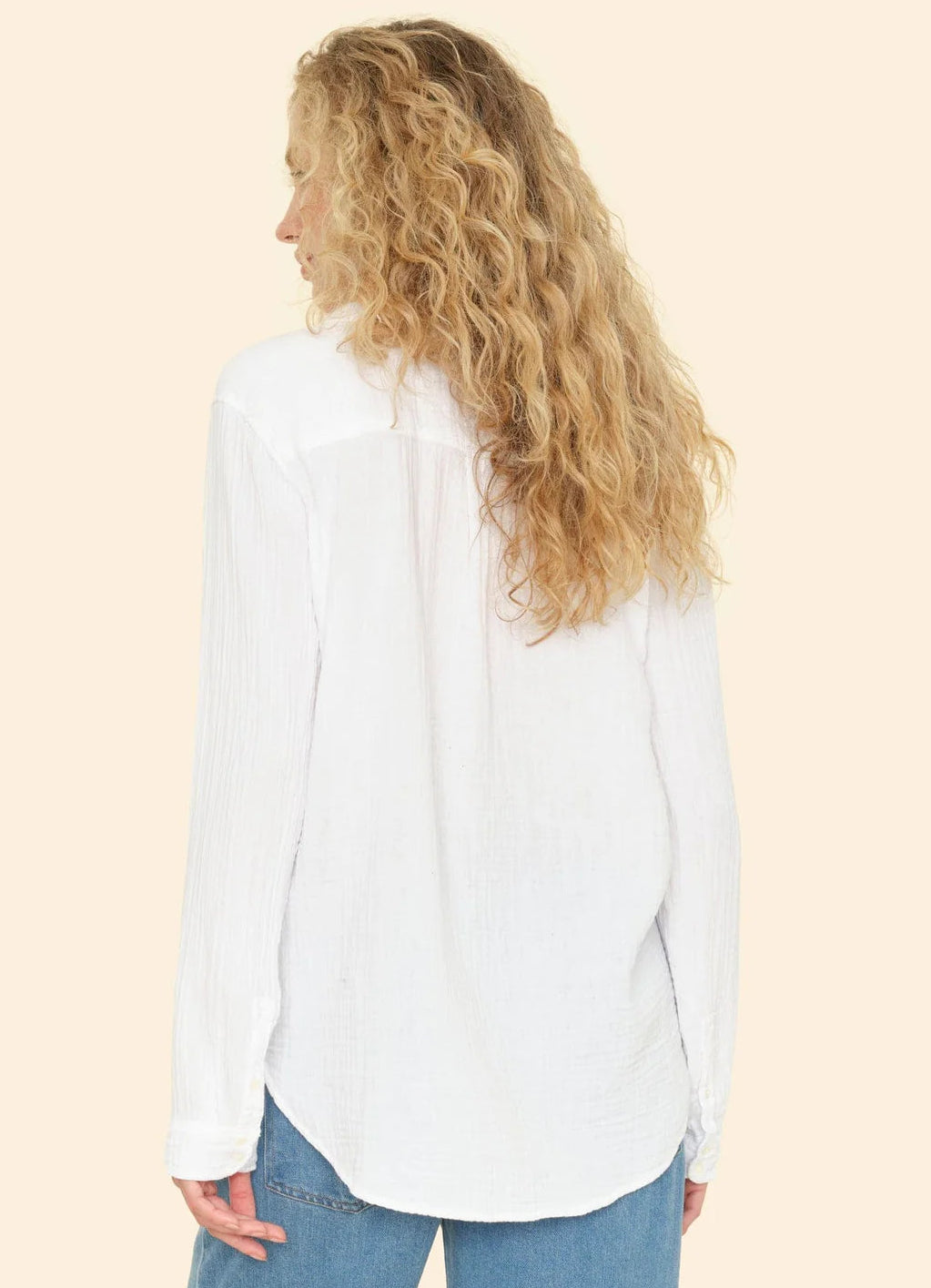 A person with long curly blonde hair, seen from behind against a beige background, wears Xirena's Scout Shirt in soft white cotton gauze paired with light blue jeans.