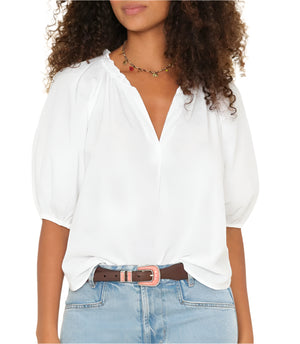 A woman wears the Xirena Jules Top—a white blouse with a ruffle neckline and short puff sleeves—styled with blue jeans, a brown belt, and a beaded necklace.