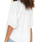 A woman with curly hair is seen from the back wearing the Xirena Jules Top, a white blouse with a ruffle neckline and puff sleeves in lightweight cotton poplin, styled with light blue jeans.