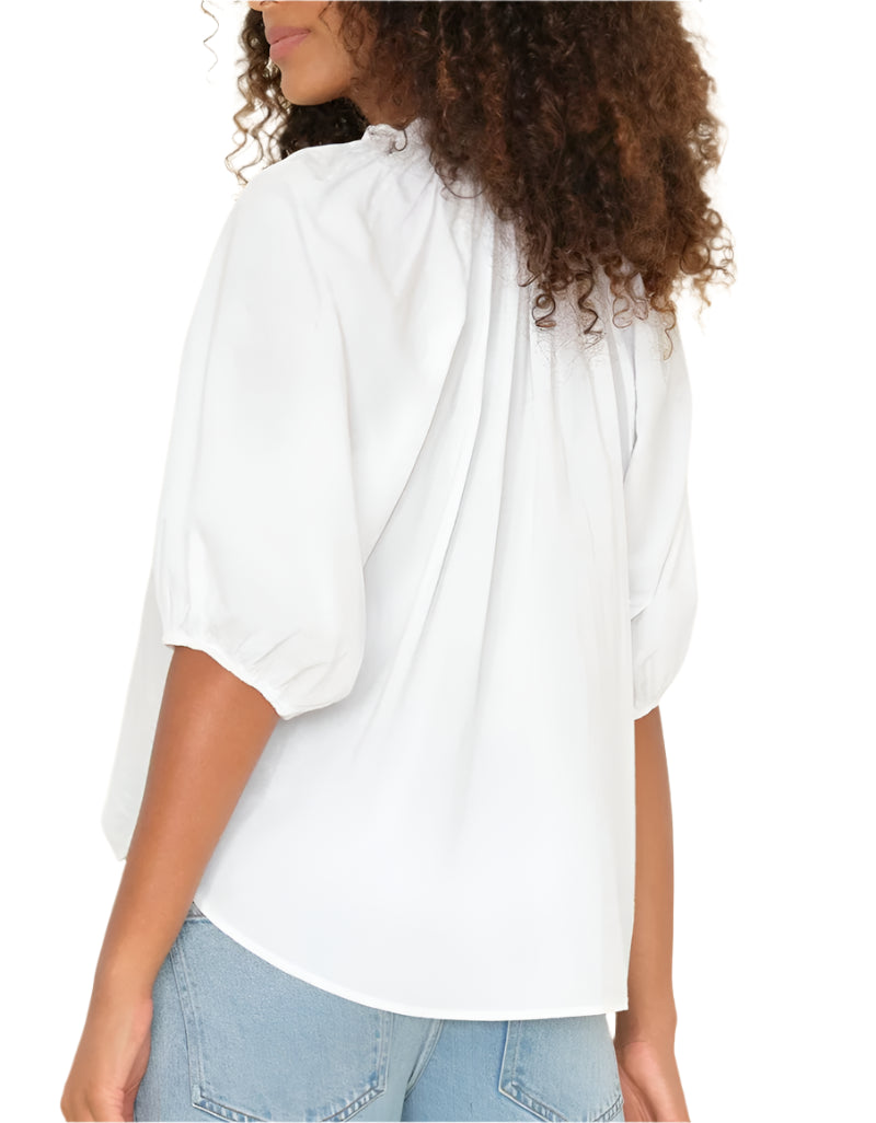 A woman with curly hair is seen from the back wearing the Xirena Jules Top, a white blouse with a ruffle neckline and puff sleeves in lightweight cotton poplin, styled with light blue jeans.