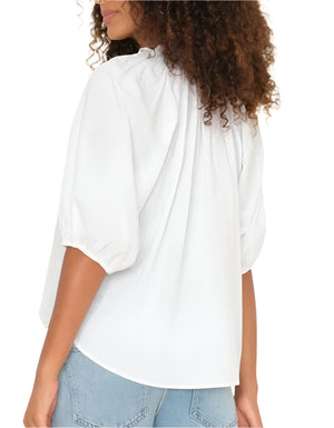 A woman with curly hair is seen from the back wearing the Xirena Jules Top, a white blouse with a ruffle neckline and puff sleeves in lightweight cotton poplin, styled with light blue jeans.